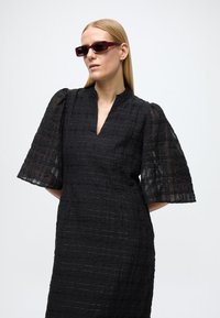 Juoda suknelė su tekstūriniu, langelių raštu, V formos iškirpte ir pūstomis, permatomomis rankovėmis. Modelis dėvi raudonas stačiakampio formos saulės akinius.