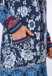 Strickpullover mit einem marineblauen Hintergrund und weißen Blumenmustern, blauen und roten Akzenten an den Bündchen und der Tasche sowie schwarzen Knöpfen.