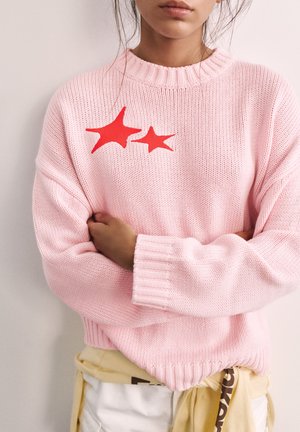 Frau, die einen rosa gestrickten Pullover mit zwei roten Sternformen auf der Brust trägt, die Arme verschränkt, und ein gelbes Kleidungsstück um die Taille gebunden hat.