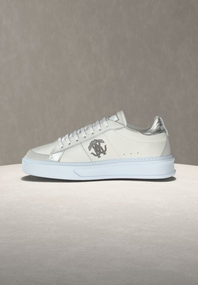 Λευκό χαμηλό αθλητικό παπούτσι με ασημένιες λεπτομέρειες και λογότυπο στο πλάι, λευκά κορδόνια και παχύ λευκό πέλμα πάνω σε ανοιχτή επιφάνεια και ουδέτερο φόντο.