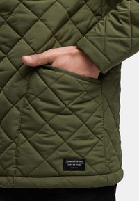 Grüne gesteppte Jacke mit seitlichem Taschen und einem gewebten Etikett am Saum, das eine glatte Textur und eine Rautenstichmuster zeigt.