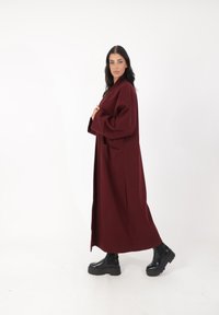 Abrigo burdeos de gran tamaño, de mangas largas, con bolsillos laterales y un diseño recto. Un par de botas negras gruesas completa el atuendo.