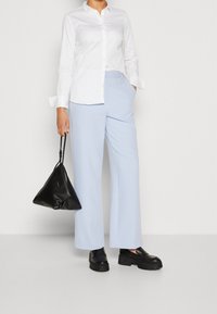 Camicia bianca a maniche lunghe con bottoni abbinata a pantaloni larghi azzurri. Tiene una borsa nera a forma di triangolo e indossa scarpe nere chunky.
