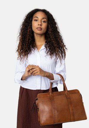 Borsa a tracolla in pelle marrone con doppio manico superiore, caratterizzata da una superficie liscia e cuciture orizzontali. Il modello tiene la borsa mentre indossa una camicia bianca e una gonna marrone.