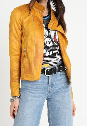 Frau trägt eine senfgelbe Lederjacke über einem gestreiften T-Shirt mit Comic-Grafik, hellblaue Jeans und einen schwarzen Nietengürtel.