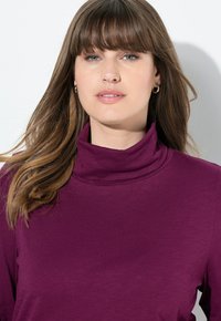 Lange mouwen, turtleneck top in dieppaars, met een zachte, gladde textuur en een minimalistisch ontwerp. Toont het gezicht en de haarstijl van het model.