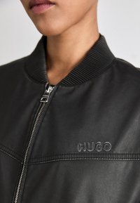 Sort læder bomberjakke med ribbestrikket krave, lynlås og præget "HUGO" logo på brystet. Glat tekstur med syns detaljer.