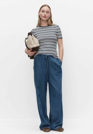 Jonge vrouw in een gestreept shirt met korte mouwen en losse blauwe jeans met hand in de zak, houdend een beige en bruine tas tegen een witte achtergrond.