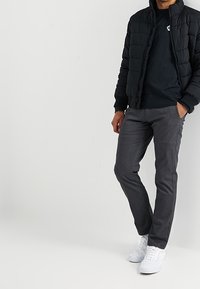 Zwarte gewatteerde jas met hoge kraag, gecombineerd met een marineblauw T-shirt en grijze broek. Het schoeisel bestaat uit witte sneakers. Overal een gladde textuur.