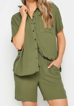 Kobieta w luźnej oliwkowozielonej koszuli na krótki rękaw z guzikami i dopasowanych szortach, z dłonią w kieszeni i luźnymi, falowanymi włosami.