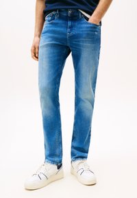 Férfi közép kék, szűk fazonú farmernadrágban és fehér sportcipőben, egyik kezét a zsebében tartva, egyszerű világos háttér előtt áll.