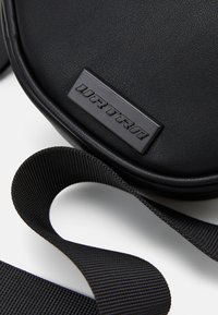 Black synthetic crossbody bag featuring a textured surface, flat strap, and a rectangular embossed logo plate.