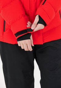 Czerwona puchowa kurtka z czarnymi akcentami, z regulowanymi mankietami na rzepy. Model ma na sobie czarne spodnie, pokazując rękę zapinającą mankiet.