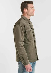 Man die naar rechts kijkt in een olijfgroene corduroy overhemd met knopen en borstzakken, genietend in een spijkerbroek, staande voor een effen achtergrond.