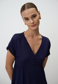 Navyblaues Kleid mit V-Ausschnitt, kurzen Ärmeln und drei dekorativen Knöpfen vorne. Hergestellt aus einem strukturierten Stoff mit fließender Silhouette.