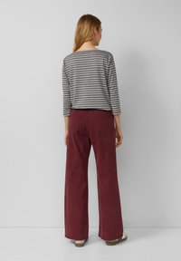 Vrouw met haar rug naar de kijker, gekleed in een grijs gestreept longsleeve shirt en hoog-waisted burgundy broek met wijde benen, gecombineerd met witte sneakers.