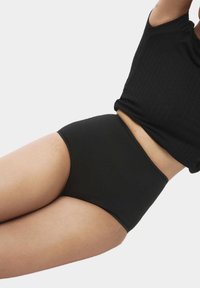 Svarta höga trosor tillverkade av mjukt tyg, med en sömlös design och mjuk elastisk midjeband, liggande mot en hudfärgad bakgrund.