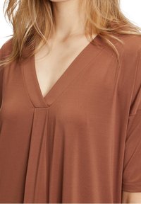 Brun V-ringad topp i mjukt tyg med plisserad design och korta ärmar. Slät textur med en avslappnad passform.