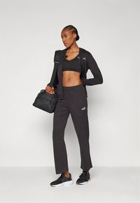 Zwart sportoutfit bestaande uit een nauwsluitende sportbeha, een jack met rits en een joggingbroek. Gemaakt van een textuurstof met een logo-accent. Draagt een zwarte tas.