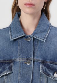 Veste en denim bleu clair avec un col, des boutons avant et deux poches poitrine. Présente des coutures visibles et une texture légèrement usée.