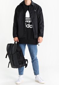 Černý batoh s texturovanou tkaninou, nastavitelnými popruhy a zaobleným klopou. Nosit jej osoba v černé bundě přes černou grafickou mikinu.