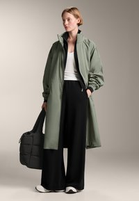 Dlouhá, olivově zelená, lehká bunda s vysokým límcem a elastickými manžetami. V kombinaci s černými kalhotami s širokými nohavicemi a bílými teniskami.