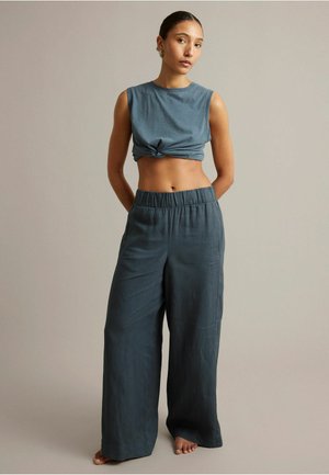 Młoda kobieta stoi boso, ubrana w bezrękawnikowy, związany krótki top i luźne, szerokie spodnie w stonowanym niebieskim kolorze na prostym tle.