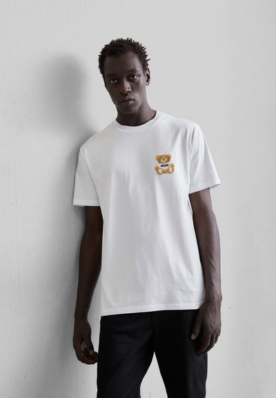 Camiseta de algodón blanca con un oso de peluche bordado en amarillo en el lado izquierdo del pecho. Simple, de manga corta y con un diseño de cuello redondo.