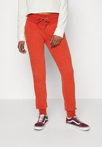 Oranžové tepláky se stahovací šňůrkou v pase, žebrovanými manžetami a bočními kapsami, v kombinaci s vínovými teniskami s bílými akcenty.