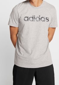 Grå bomullströja med korta ärmar som har en tydlig, mörkgrå Adidas-logotyp på bröstet i ett camouflagemönster.