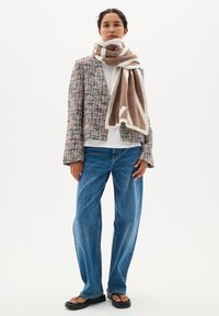 Sacou din tweed țesut multicolor, pulover alb cu guler înalt, blugi casual albaștri, fular maro cu motive și margini albe, sandale plate negre.