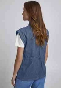 Jeansowa kamizelka z dopasowanym wcięciem w talii, bez rękawów, uszyta z textured blue materiału. Nosi się ją na białej koszulce.