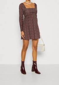 Robe à manches longues florale avec un encolure carrée, fond noir et fleurs rouges. Portée avec des bottines bordeaux et un sac à main blanc.