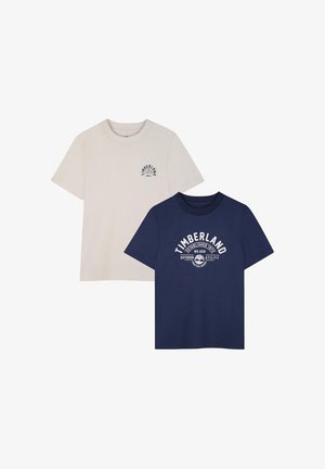 Due magliette Timberland a maniche corte con scollo rotondo; una beige con piccolo logo, una blu navy con grande logo bianco e testo sul davanti.