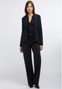 Schwarzer taillierter Blazer und passendes Hemd mit Knöpfen, kombiniert mit weit geschnittenen schwarzen Hosen. Glattes Material und strukturiertes Design.