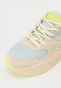 Sneaker with a light blue mesh upper, beige suede accents, and vibrant yellow laces. Textured sole in pastel pink with a green detail.
