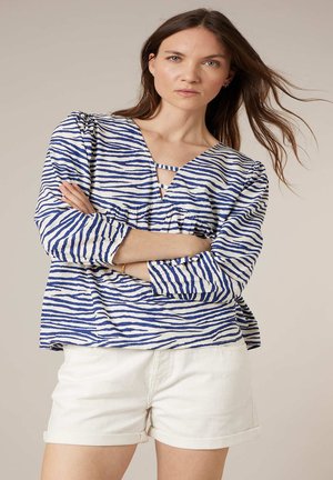 Femme aux longs cheveux bruns portant un chemisier à rayures bleues et blanches et un short blanc, debout les bras croisés devant un fond neutre.