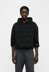Μαύρο βελούδινο φούτερ με υφή "Air Jordan", που διαθέτει τσέπη καγκουρό και μικρό λογότυπο στο μανίκι. Συνδυασμένο με γκρι παντελόνι.