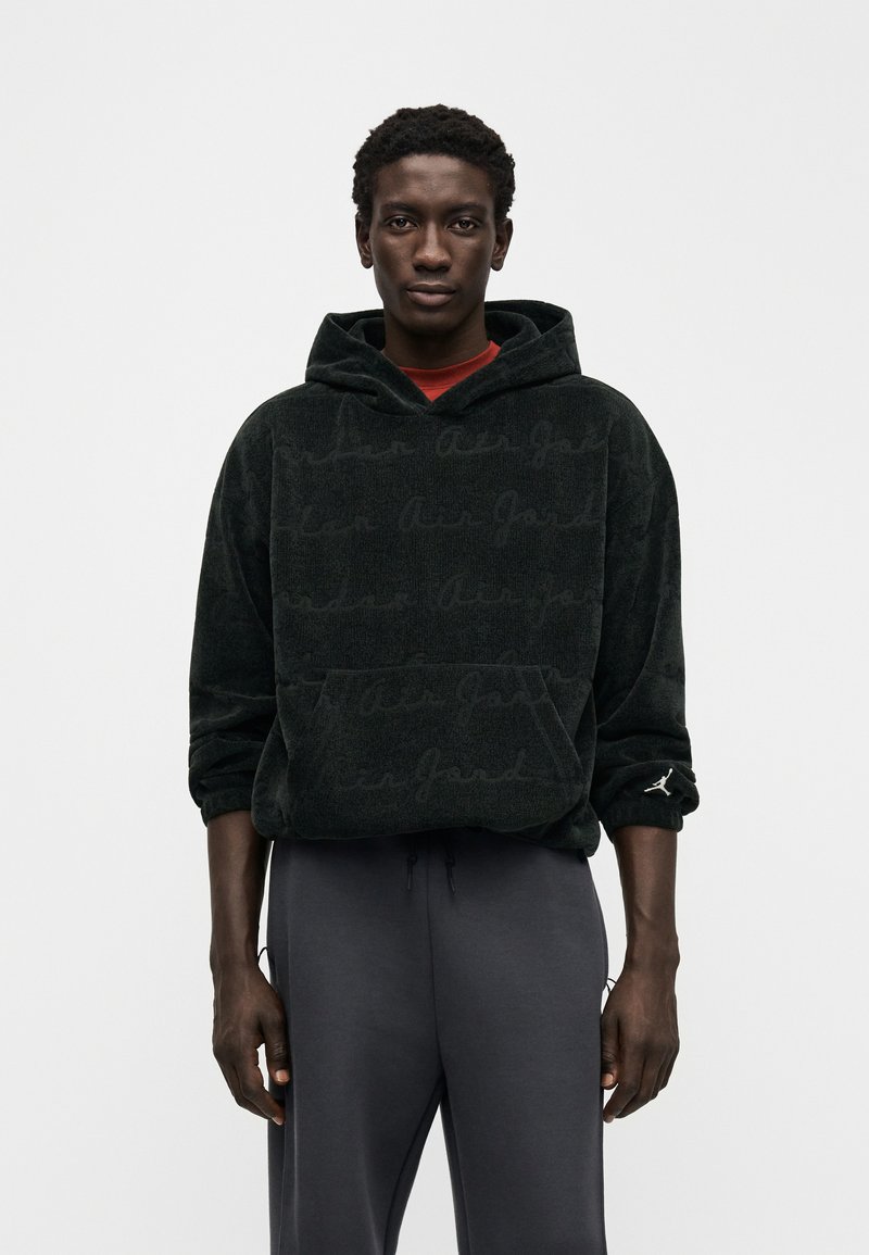 Μαύρο βελούδινο φούτερ με υφή "Air Jordan", που διαθέτει τσέπη καγκουρό και μικρό λογότυπο στο μανίκι. Συνδυασμένο με γκρι παντελόνι.