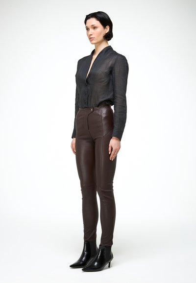 Kobieta o krótkich ciemnych włosach, ubrana w czarną przejrzystą koszulę na guziki, brązowe obcisłe skórzane spodnie i czarne botki, stojąca na białym tle.