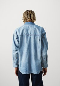 Jasnoniebieska koszula z denimu z długimi rękawami, karo na plecach i mankietami na guziki. Zaokrąglony dół i miękka faktura, z delikatnym detalem przeszycia.