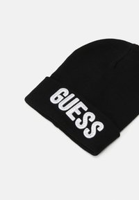 Berretto a maglia nero con risvolto, caratterizzato dalla parola "GUESS" ricamata in lettere bianche, adornato con borchie decorative.