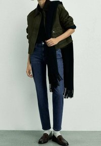 Veste vert olive avec col marron, écharpe noire, jean bleu foncé ajusté, chaussettes blanches et mocassins marron. Design simple et structuré.