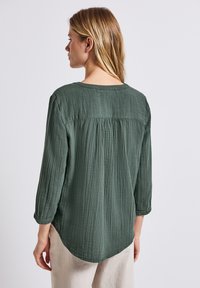 B blouse verde, con texture, dalla scollatura rotonda e maniche a tre quarti; presenta una parte posteriore arricciata e un'orlo curvo più lungo.