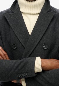 Dunkelgrauer Fischgrätenmantel mit doppelreihigem Design und schwarzen Knöpfen. Cremefarbener, gerippter Rollkragenpullover ist darunter zu sehen und zeigt einen kontrastierenden Stoff.