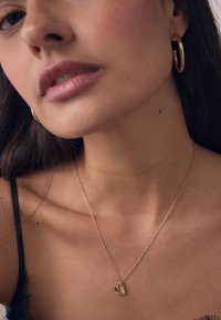 Close-up of a woman wearing a gold chain necklace with charms, gold hoop earrings, and a black lace strap top.