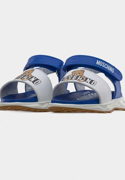 Μπλε και λευκά σανδάλια για νήπια με εικονογράφηση αρκουδάκι, ιμάντες με βέλκρο και την επωνυμία "Moschino" στους ιμάντες στους αστραγάλους.