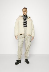 Fuzzy krämfärgad och persikofärgad jacka, svart och vit randig polotröja, beige cargobyxor med sidofickor och svarta sneakers.