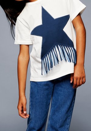 Niño con camisa blanca de manga corta con una gran estrella azul marino y detalles de flecos, combinado con jeans de mezclilla azul sobre un fondo gris.