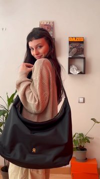 Νεαρή γυναίκα με μπεζ πουλόβερ κρατά μεγάλη μαύρη τσάντα σε εσωτερικό χώρο με φυτά, πορτοκαλί κουτιά και ραφιέρες στον τοίχο με περιοδικά.